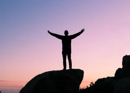 Person standing triumphantly on top of a rocky peak, arms raised towards the sky in a gesture of achievement and self-empowerment, with a backdrop of a clear horizon symbolizing clarity and personal growth.